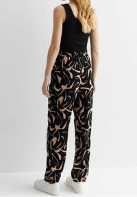 Vrouw draagt een zwarte mouwloze top en losse zwarte broek met een beige abstract patroon, gecombineerd met witte sneakers, en staat met haar rug naar de camera.
