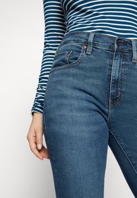 Modré džíny s vysokým pasem mají klasický design s pěti kapsami, prošité detaily a hladký povrch, vedle kterých odpočívá ruka modelky.