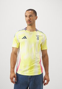 Gele voetbalshirt met een mesh-textuur, met blauwe accenten, drie sterren en roze zijpanelen. Korte mouwen en een ronde halsontwerp.