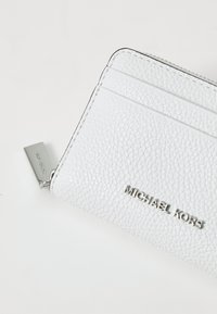 Valge tekstuuriga nahast kaardihoidja, millel on mitmeid kaardisahtleid, hõbedane riistvara ja bränditud metallmärk, millel on kiri "MICHAEL KORS."
