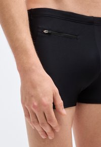 Μαύρα μαγιό σορτς με λεία υφή, που διαθέτουν τσέπη με φερμουάρ στο πίσω μέρος και εφαρμοστή σχεδίαση στη μέση και στους μηρούς.