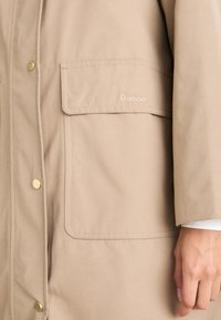 Manteau Barbour beige avec grande poche avant et boutons-pression dorés, montrant partiellement la main et la manche d'une personne.
