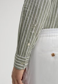 Grün-weiß gestreiftes Leinenhemd mit Knopfmanschetten, über weißen Hosen getragen, die einen einzelnen Holzknopf an der Gesäßtasche haben.