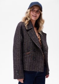 Kobieta z blond lokami, w granatowej czapce, brązowej marynarce w wzory i ciemnoniebieskich dżinsach, spogląda w bok z delikatnym uśmiechem.