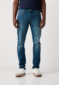 Jeans de denim azul con un ajuste ajustado, textura desgastada y costuras naranjas. Combinados con zapatillas blancas que tienen detalles en gris claro.