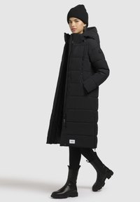 Lungo cappotto imbottito nero con cappuccio, dotato di zip anteriore, tasche laterali e un patch con logo. Indossato con pantaloni neri aderenti e stivaletti alla caviglia.