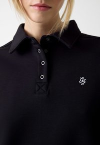 Zwart poloshirt met een klassieke kraag, voorzien van een knoopsluiting met vier zilverkleurige knopen en een wit geborduurd logo op de borst.