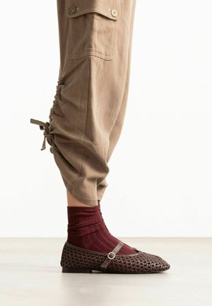 Chaussure plate Mary Jane tissée marron avec boucle portée sur des chaussettes bordeaux côtelées et un pantalon beige raccourci avec poches boutonnées et détail noué.