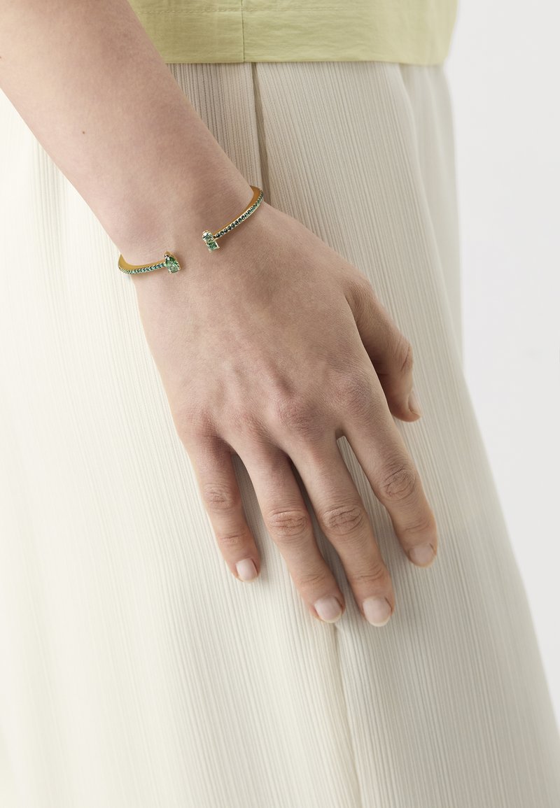 Gouden armband met groene edelstenen, gekenmerkt door twee accenten, gedragen om een hand die rust op een lichte geplooide stof. Glad oppervlakte en eenvoudig ontwerp.