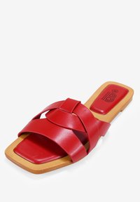Mules en cuir rouge avec des brides tressées, semelle plate et bout carré. La semelle est beige avec une légère texture et un détail de marque.