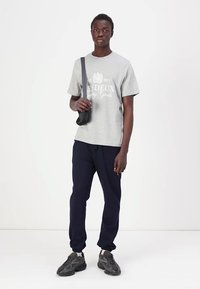 Tricou din bumbac gri cu text alb imprimat, asociat cu pantaloni joggeri bleumarin. Modelul poartă o geantă mică neagră de umăr și adidași gri.