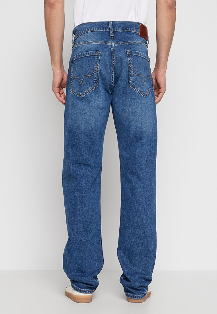 Ein Mann trägt gerade blaue Jeans und weiße Sneaker, von hinten vor einem einfarbigen, hellen Hintergrund gezeigt.