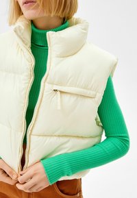 Cream puffer vest with high collar and front zip, featuring a small zippered pocket. Worn over a green ribbed turtleneck.