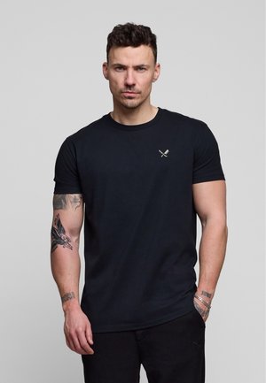 Mann mit kurzen dunklen Haaren und Tattoos trägt ein schwarzes T-Shirt mit kleinem gekreuztem Besteck-Logo auf der Brust, steht vor grauem Hintergrund.
