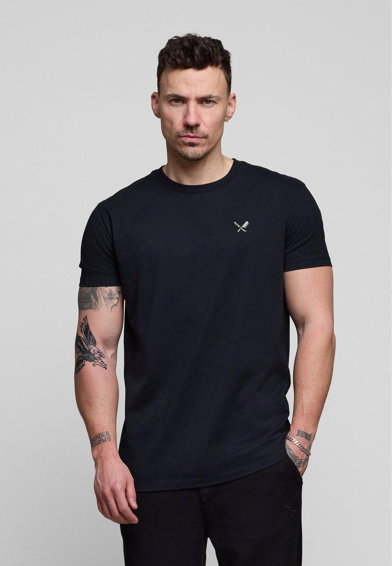 Homme aux cheveux courts et foncés et tatouages portant un T-shirt noir avec un petit logo d'ustensiles croisés sur la poitrine, debout devant un fond gris.