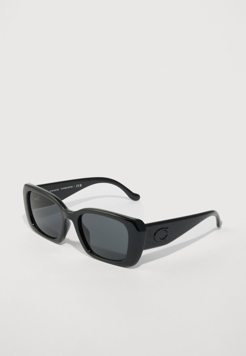 Schwarze rechteckige Coach-Sonnenbrille mit dunklen Gläsern und einem subtilen "C"-Logo am Bügel, schräg auf einem weißen Hintergrund dargestellt.
