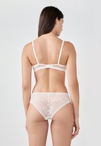 Koronkowy zestaw biustonosza i majtek w jasnym kremowym kolorze. Charakteryzuje się kwiatowymi wzorami, regulowanymi ramiączkami i falbaniastymi brzegami. Miękka, teksturowana tkanina.