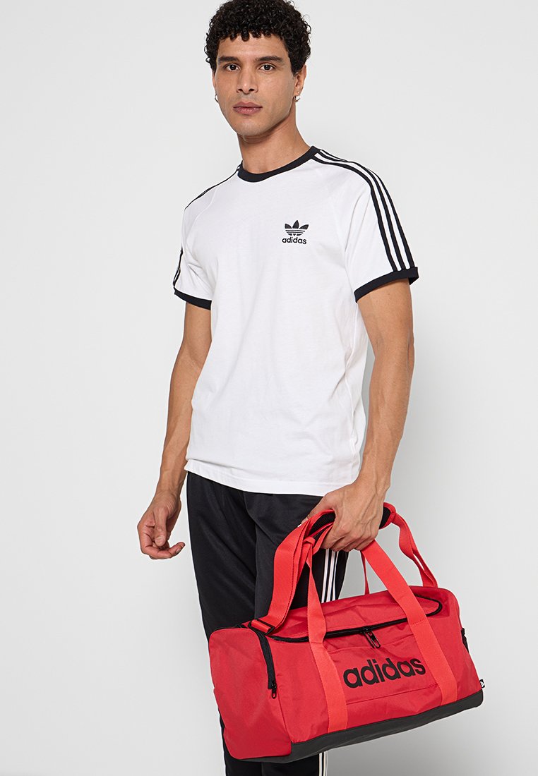 Bolsa de deporte roja con detalles en negro, que cuenta con dos asas, un cierre de cremallera y el logo de Adidas exhibido de manera prominente. Acabado de tela texturizada.
