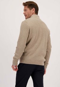 Man in een beige textuur gebreide trui en donkere broek, iets gedraaid met de rug en zijprofiel tegen een effen achtergrond.