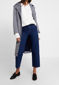 Šedý dlouhý kabát přes bílý žebrovaný svetr, doplněný o tmavě modré džíny s vysokým pasem a černé mokasíny. Kabát má strukturovaný střih.