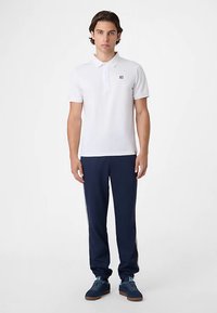 Weißes Poloshirt mit kurzen Ärmeln und kleinem Logo, kombiniert mit marineblauen Jogginghosen mit weißen Seitenstreifen und strukturierten Sneakern.
