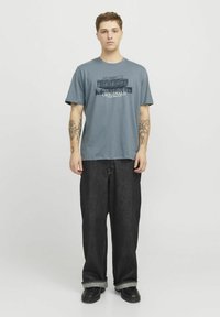 Tricou gri din bumbac cu mâneci scurte și un imprimeu grafic cu textul „ORIGINALS.” Asortat cu blugi din denim închis, cu croială lejeră, și pantofi negri.