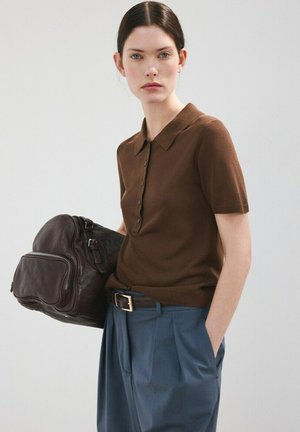 Kvinde iført brun kortærmet poloshirt og blå bukser, der holder en mørkebrun lædertaske med den ene hånd i lommen.