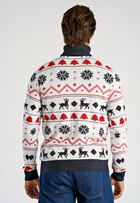 Pull de vacances tricoté avec un fond blanc orné de motifs noirs, rouges et verts représentant des rennes, des flocons de neige et des formes géométriques.
