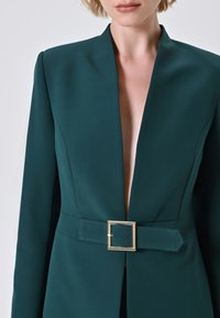 Blazer teal realizzato in tessuto liscio con scollatura profonda, maniche lunghe e una fibbia rettangolare dorata in vita.