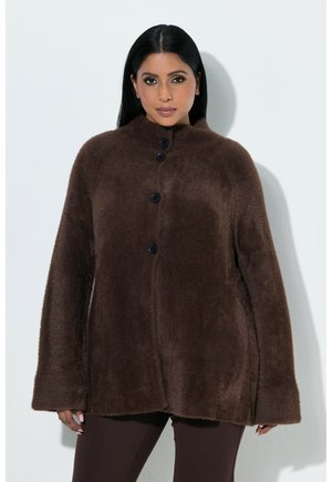 Vrouw met lang zwart haar draagt een donkerbruin, pluizig knoopjas en donkere broek, staand tegen een witte achtergrond.