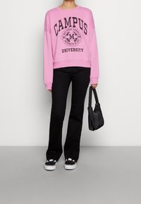 Sudadera rosa con el texto "CAMPUS UNIVERSITY", pantalones de campana negros y bolso negro. Contra un fondo liso.