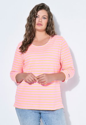 Frau mit langen gewellten Haaren, die ein rosa und gelb gestreiftes Langarmshirt und hellblaue Jeans trägt, steht vor einem weißen Hintergrund.