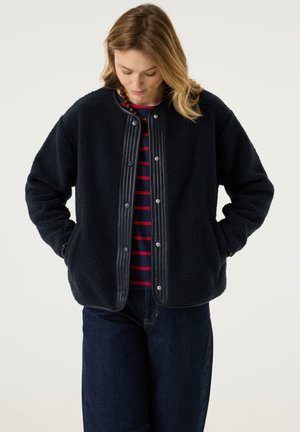 Frau steht mit den Händen in den Taschen, trägt eine marineblaue Fleecejacke über einem rot-blau gestreiften Shirt und blickt nach unten vor einem schlichten Hintergrund.
