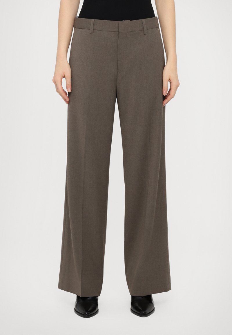 Pantalon gris à jambe large en tissu lisse et texturé, doté d'une taille classique et de passants de ceinture, porté avec des chaussures noires pointues.