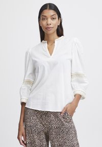 Blouse en lin blanc avec des manches bouffantes, décolleté en V et accents texturés. Portée avec un pantalon à imprimé léopard marron.