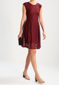 Vestido de encaje burdeos con patrón de ondas, mangas cortas y falda acampanada. Combinado con zapatos de tacón bloque en tono nude y un pequeño bolso negro.