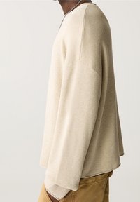 Maglietta a maniche lunghe color beige chiaro con texture a coste, scollatura arrotondata e vestibilità leggermente oversize. La vista laterale mostra una silhouette rilassata.