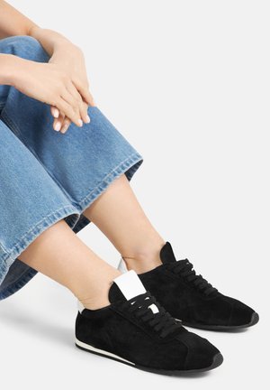 Osoba w czarnych zamszowych tenisówkach z białym językiem i detalem na pięcie, siedząca ze skrzyżowanymi nogami, ubrana w krótkie niebieskie dżinsy.