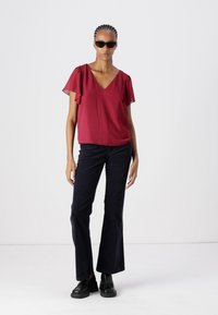 Burgundy chiffon top with short flutter sleeves, paired with dark flared jeans and black chunky shoes. Model wearing oversized sunglasses.