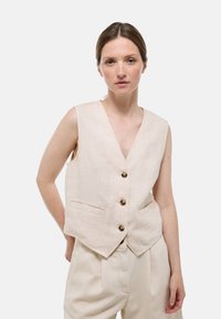 Gilet en lin beige avec un col en V, cinq boutons et deux poches avant. Le design est sans manches et ajusté avec un ourlet légèrement incurvé.