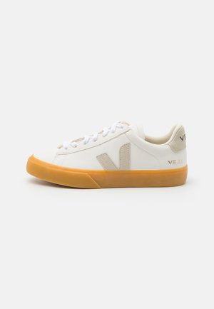 Zapatilla blanca de perfil bajo con logo "V" y lengüeta trasera en ante beige, suela de goma marrón y cordones blancos sobre fondo claro liso.