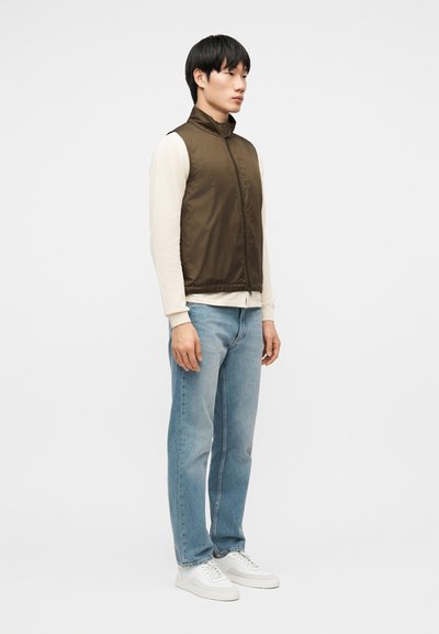 Νεαρός άνδρας όρθιος, φοράει καφέ αμάνικο γιλέκο πάνω από κρεμ μακρυμάνικο πουκάμισο, ανοιχτό μπλε τζιν και λευκά αθλητικά παπούτσια, κοιτάζοντας δεξιά.