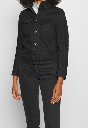 Frau trägt eine eng anliegende schwarze Jeansjacke mit Knopfverschluss und schwarze Jeans, steht vor einem schlichten, hellen Hintergrund.