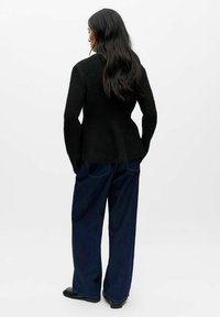 Musta, teksturoitu neulejakku leveällä helmaosalla, yllään tummansiniset leveälahkeiset farkut. Pitkät, suorat hiukset näkyvissä. Minimalistinen tausta.