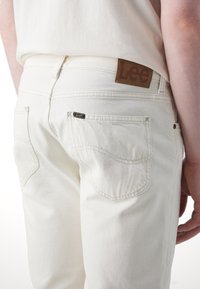 Blugi din denim alb cu o formă ajustată, având un design cu buzunare cusute, o eticheta din piele maro la talie și detalii de cusătură vizibile.