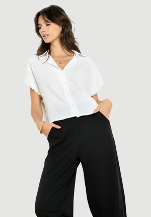 Vrouw die een losse witte korte mouw blouse met knopen en brede zwarte broek draagt, staand met handen in de zakken tegen een effen achtergrond.