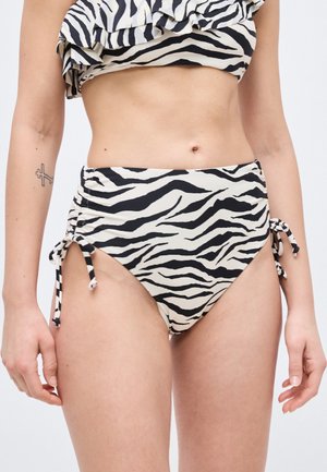 Frau trägt schwarze und weiße Bikinihose mit Zebramuster, hohen Bund und seitlichen Bändern sowie ein passendes, gerafftes Oberteil.