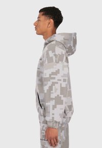 Tânăr îmbrăcat într-un hanorac cu camuflaj în nuanțe de gri și alb, împreună cu pantaloni asortați, stând în profil pe un fundal simplu.