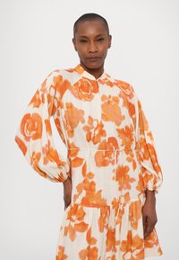 Blumenkleid aus leichtem Stoff, mit orangefarbenen und pfirsichfarbenen Blumenmustern auf einem cremefarbenen Hintergrund, langen Ärmeln und einem gerafften Saum.