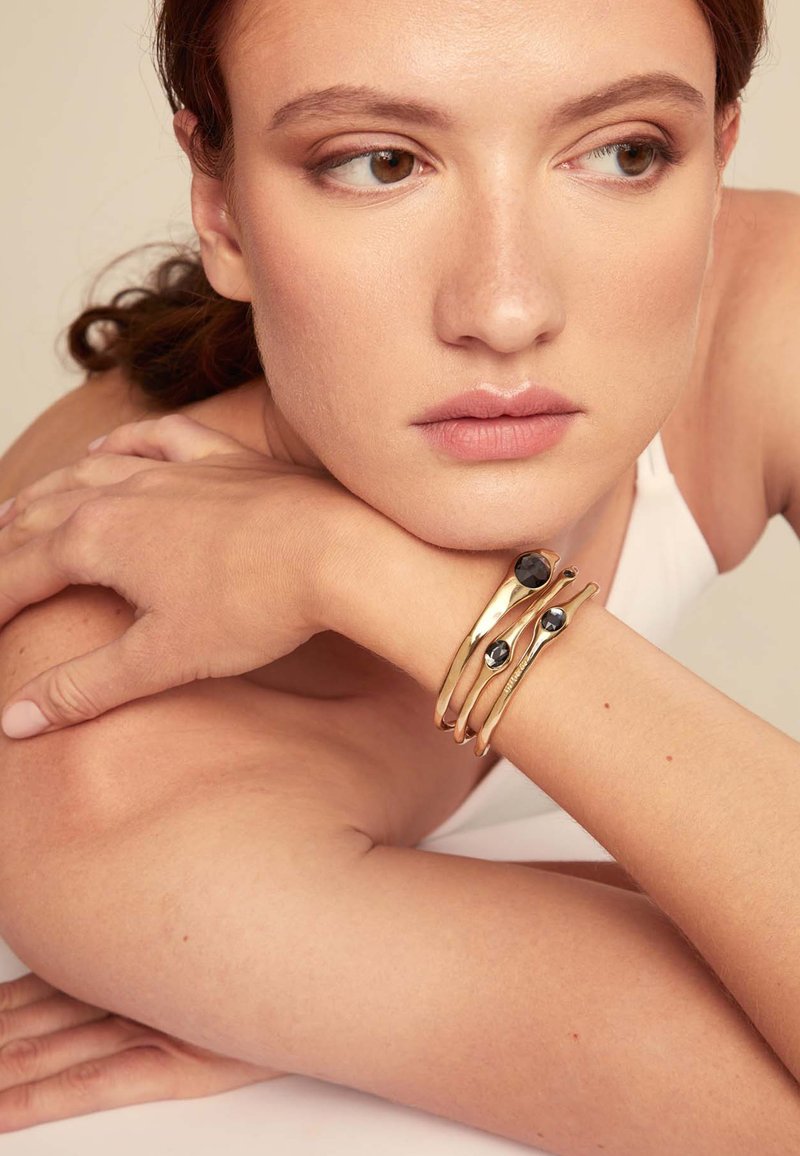 Trois bracelets en métal doré avec des accents noirs, présentant des courbes douces et un design épuré, portés sur un poignet de teint naturel.